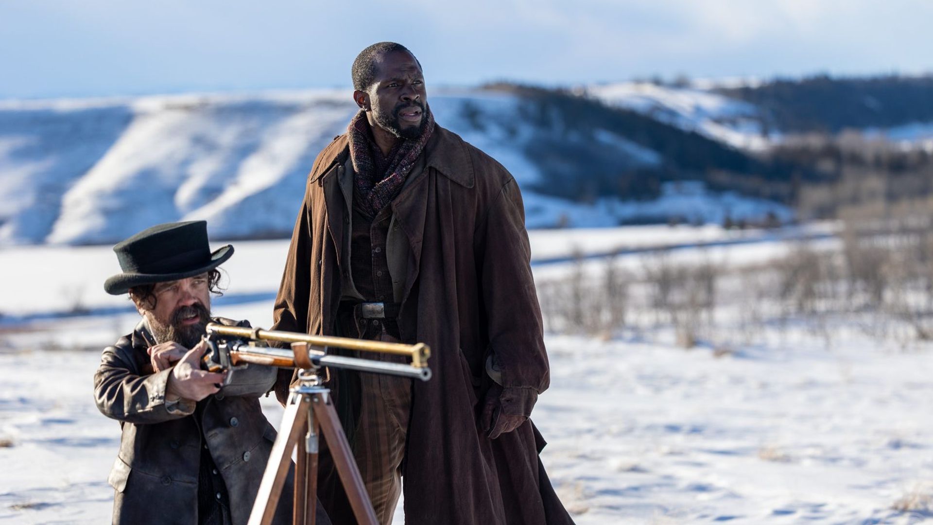 Питер Динклэйдж и Гбенга Акиннагбе стоят на снегу, один целится из винтовки с треногой, другой наблюдает вдаль
