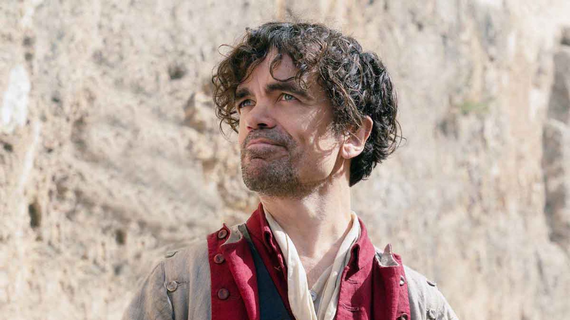 Питер Динклэйдж в историческом костюме с красным камзолом держит письмо и смотрит вверх с лёгкой улыбкой