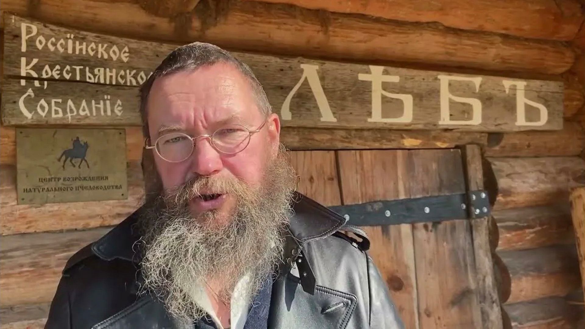 Пожилой мужчина с кудрявой седой бородой и в очках, сзади него деревянная изба